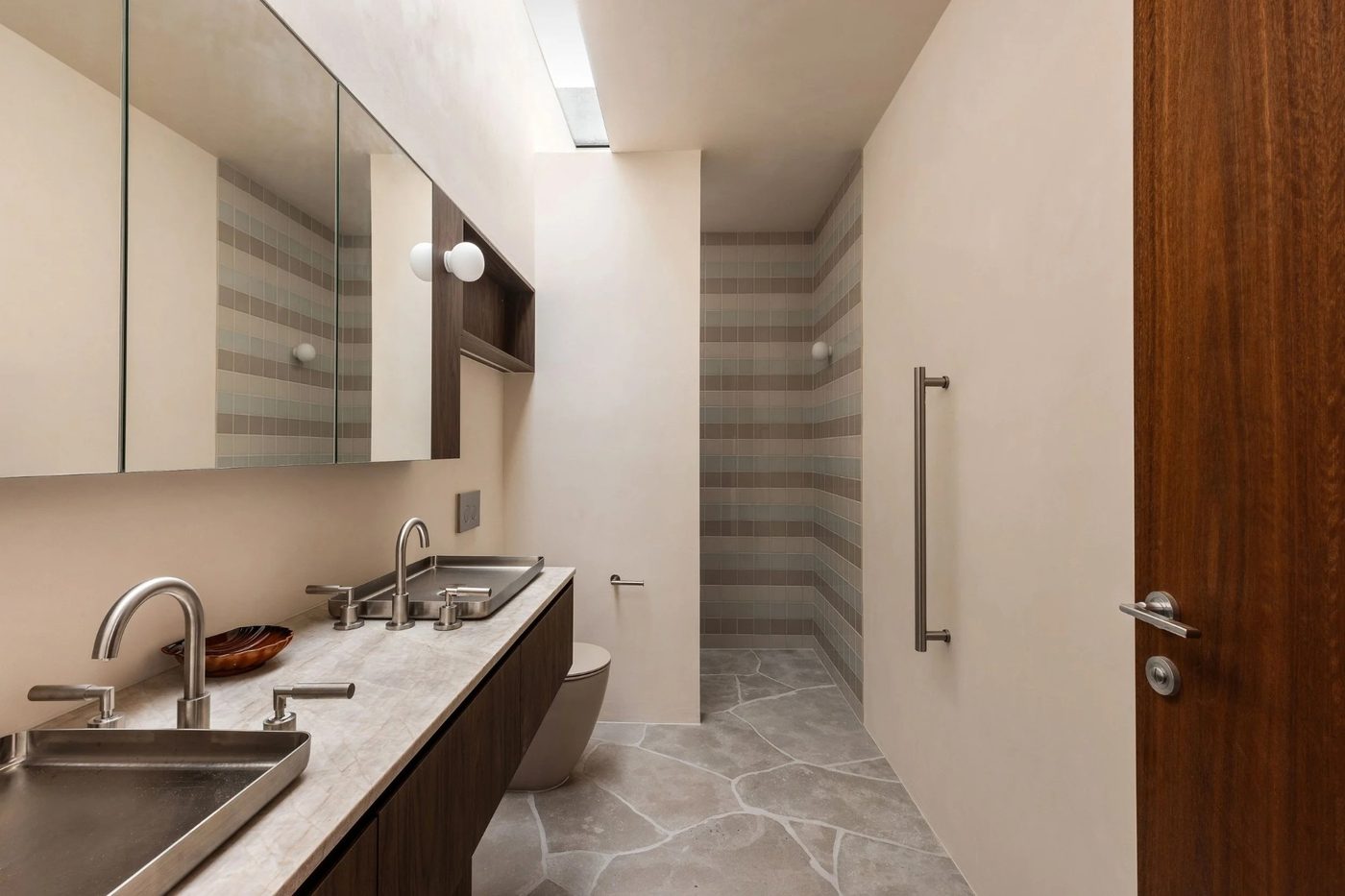 Morada Build bathroom with stone-topped walnut vanity, twin stainless basins and gooseneck tapware, mirrored cabinetry and crazy-paved stone floor leading to a tiled shower
